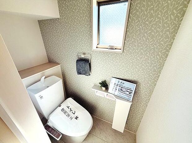 窓付きで換気しやすいトイレです。グレージュ系のアクセントクロスが落ち着いた印象を与えます。手洗いスペースと収納棚もあり使い勝手にも配慮されています。ペーパーや小物もすっきり置ける設計です。