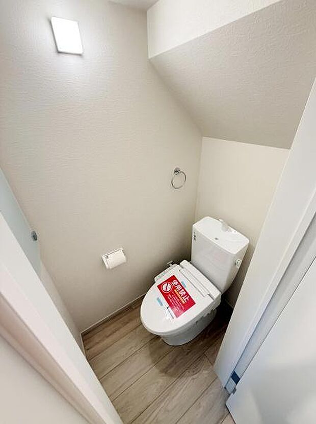 シンプルで清潔感のあるトイレです。壁付けの照明が明るさを確保しています。床は木目調で温かみがあります。ペーパーホルダーやタオルリングも設置されています。