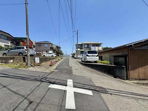 現地前面道路は幅があり見通しの良い直線道路です。周辺には住宅が並んでおり落ち着いた雰囲気が感じられます。