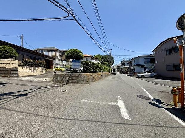 広々とした前面道路が特徴的な現地写真です。周囲には住宅が建ち並び落ち着いた雰囲気が感じられます。電線や標識も整然と配置されています。