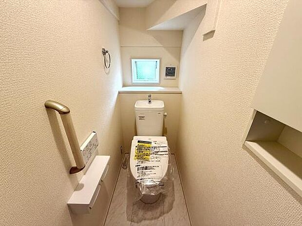 明るい色合いの内装が印象的なトイレです。窓があり換気に便利です。手すり付きで使いやすさが考慮されています。収納スペースも確保されています。清潔感のある空間です。