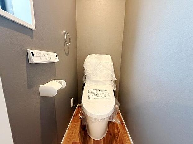 落ち着いた色合いの壁と木目調の床が調和した空間です。新しさを感じるトイレが設置されています。ペーパーホルダーやリモコンも使いやすい位置に配置されています。清潔感が保たれています。