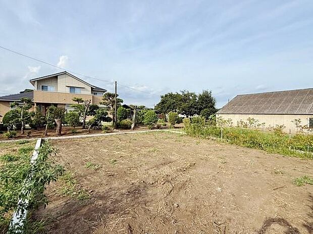 周辺には住宅が建ち並び落ち着いた雰囲気の現地土地写真です。青空が広がり開放感があります。平坦な土地で扱いやすく多様なプランが検討しやすい形状です。前面道路への接続も便利です。