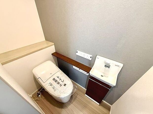 落ち着いた色合いの壁とフローリングが調和したトイレ空間です。手洗いカウンターと収納スペースもあり機能的なレイアウトとなっています。清潔感のある設計で日々の利用もしやすいです。