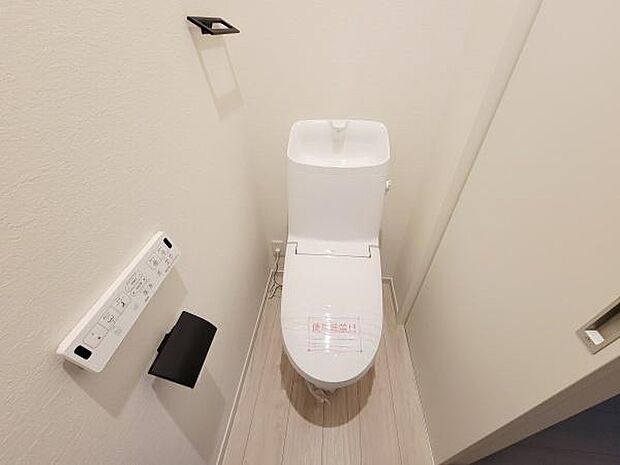 清潔感のあるトイレ空間です。温水洗浄便座が設置されています。壁付けのペーパーホルダーと操作パネルが使いやすい位置に配置されています。床は木目調で落ち着いた印象です。