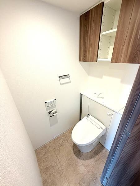清潔感のあるトイレ空間です。収納棚が設置されており整理整頓がしやすい仕様です。明るい色合いで開放感を感じられます。温水洗浄便座も付いています。ドアや棚などの木目調がアクセントとなっています。