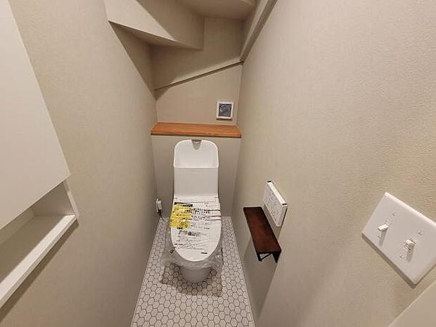 シンプルなデザインのトイレ空間です。壁面には飾り棚があり小物の設置に便利です。六角形タイルの床がアクセントとなっています。照明スイッチが手元にあり操作も容易です。