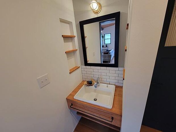木目調のカウンターと白いタイルの組み合わせがシンプルな印象の洗面台です。壁面の小物棚がアクセントとなり機能的な空間を演出しています。黒縁のミラーが空間を引き締めています。