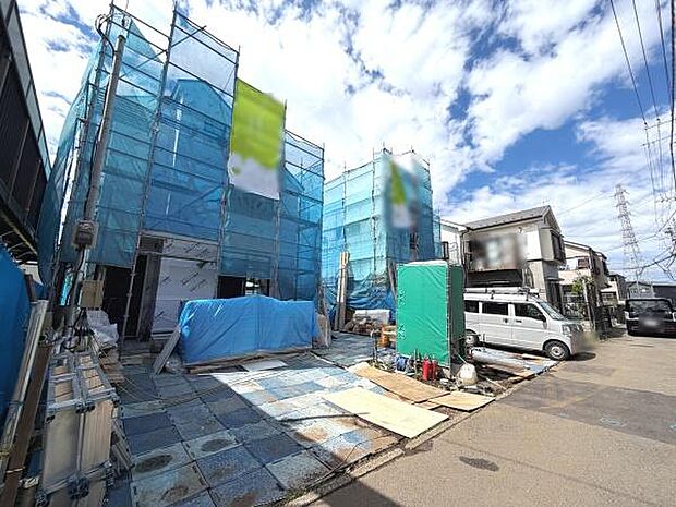 青空の下建築中の建物が並ぶ現場です。足場や養生シートがしっかり設置されており工事の進捗が感じられます。周辺は住宅が立ち並ぶ落ち着いた環境です。車の駐車スペースもしっかり確保されています。