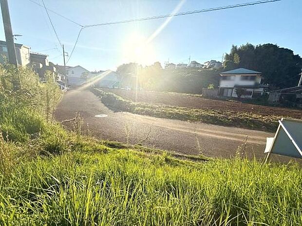 前面道路が広がり日当たりも感じられる現地写真です。周囲は自然が多く穏やかな街並みが広がっています。