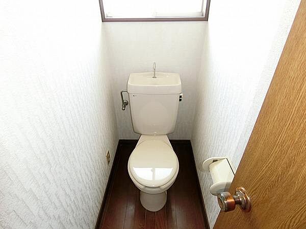 シンプルなデザインのトイレです。上部に窓があり明るい空間を実現しています。床は木目調で清潔感があります。ペーパーホルダーも設置済みで使いやすさに配慮されています。扉を開けやすい取っ手が付いています。