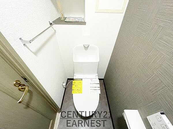 シンプルなデザインで清潔感を感じるトイレです。ペーパーホルダーや棚が設置され使い勝手も良好です。壁面のクロスと床材が調和した空間となっています。収納スペースも備えられており機能面も配慮されています。