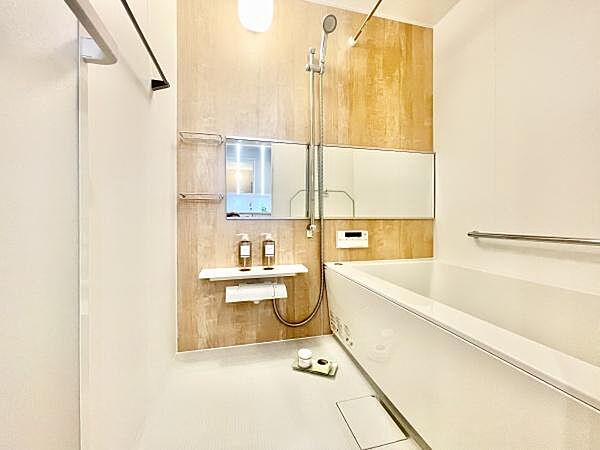 明るい照明と木目調の壁が魅力的なバスルームです。大きな鏡が空間を広く感じさせます。バスタブには手すりが設置されており使いやすさが感じられます。収納スペースも確保されており日常使いに便利です。