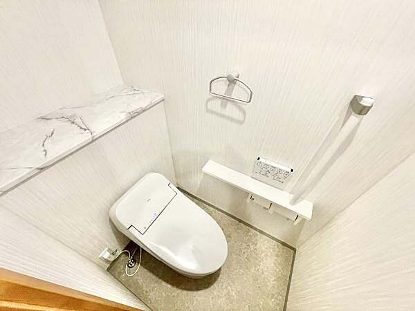 清潔感のある白を基調としたトイレです。手すりやペーパーホルダーが設置され使いやすさにも配慮されています。奥には棚スペースがあり収納に便利な設計です。壁面がシンプルなため空間がすっきりと感じられます。