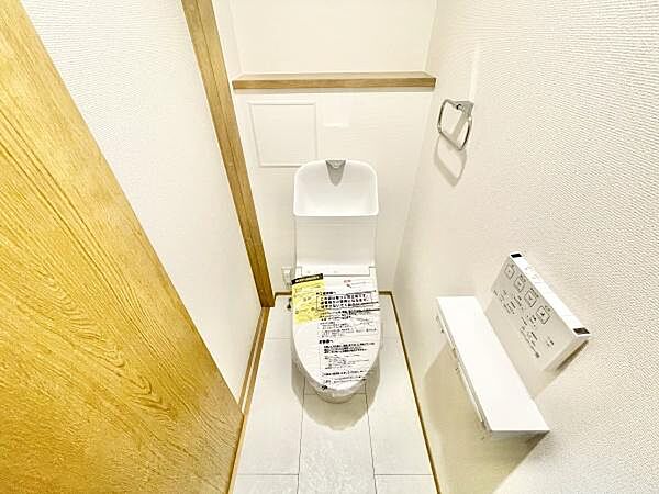シンプルで清潔感のあるトイレ空間です。収納棚が備え付けられており小物の整理もしやすくなっています。壁付けのペーパーホルダーや操作パネルが使いやすい位置に配置されています。
