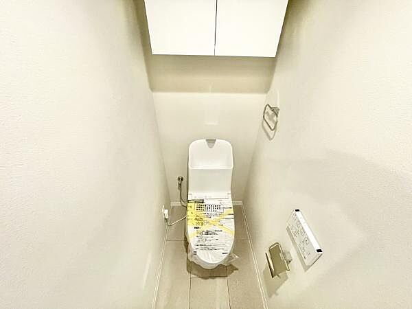 シンプルなデザインのトイレです。上部収納棚が備え付けられており使い勝手が良好です。壁面にはペーパーホルダーやタオル掛けが設置されています。清潔感のある空間です。