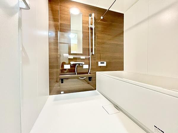 木目調のアクセントパネルが印象的なバスルームです。広々とした空間で清潔感のある白い床と壁が特徴です。浴槽はゆったりとしたサイズで毎日のリラックスタイムに適しています。