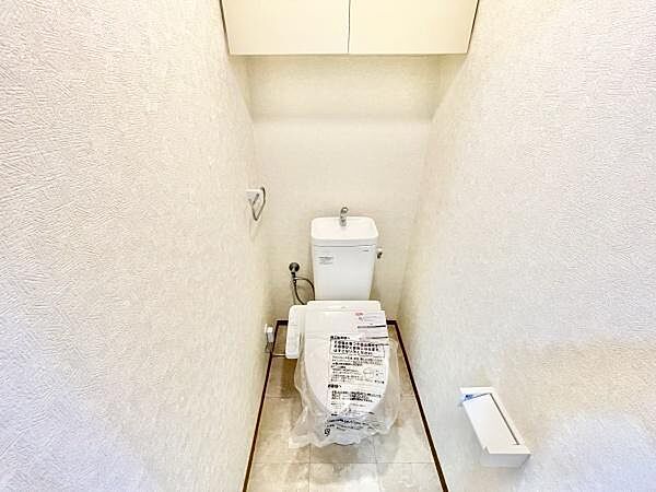 清潔感のあるシンプルなトイレ空間です。上部収納棚が設けられており小物の整理にも役立ちます。壁紙は明るい色合いで空間に広がりを感じます。ペーパーホルダーも使いやすい位置に設置されています。