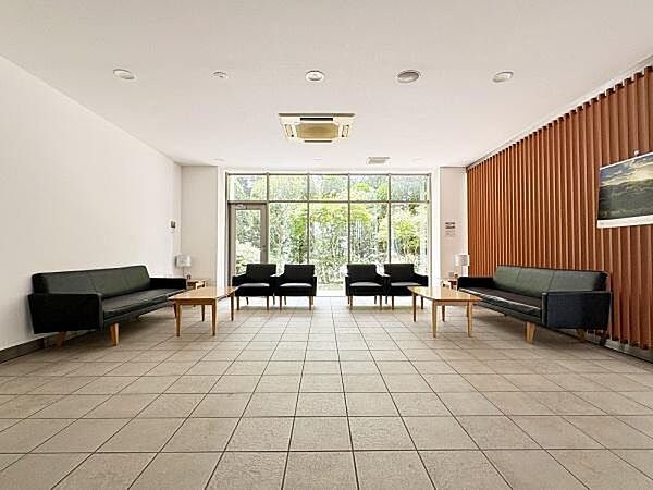 木製ルーバーの壁面と窓外の植栽が落ち着いた趣を添える共用ラウンジです。足元には上品なタイルが広がり、テーブルを囲むソファ席で、外の木漏れ日を眺めながら待ち合わせや休息のひとときを過ごせます。