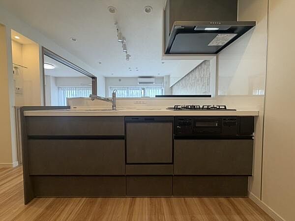 木目調の床とダークカラーのキッチンが調和したデザインです。天板が広く作業スペースがしっかりと確保されています。ガスコンロや換気設備も整っており機能性も感じられます。