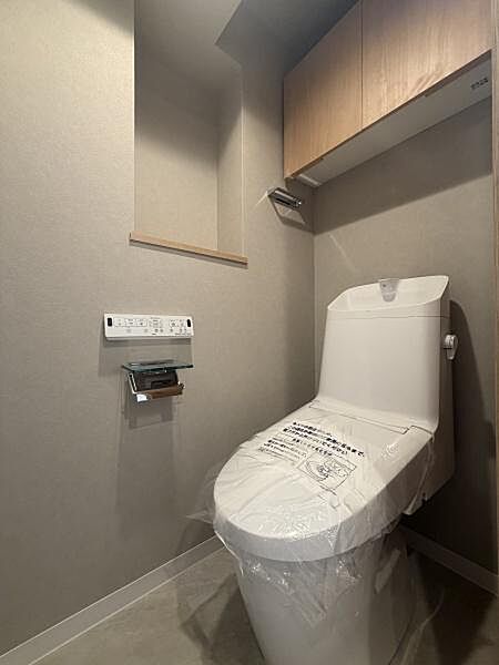 清潔感のあるトイレ空間です。壁面には収納棚が設けられており利便性が考慮されています。温水洗浄便座のリモコンも設置されています。明るい内装が印象的です。全体的にすっきりとした印象です。