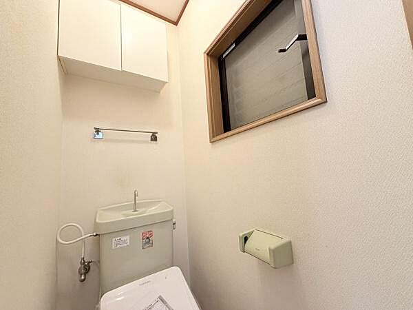 上部に収納棚が設けられておりトイレットペーパーなどの整理に便利です。換気に役立つ窓が設置されているため明るさも確保されています。壁に備え付けのタオル掛けもあり機能的な空間です。