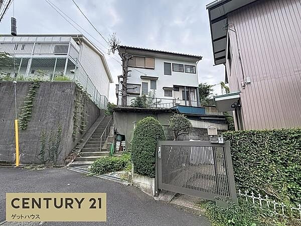 落ち着いた住宅地に位置する現地写真です。前面道路は車の通行も視認しやすく安心感のある環境です。玄関前の門扉や植栽が整えられており敷地との境界も明確です。周囲の建物や道路の様子がひと目で分かる一枚です。