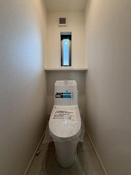 コンパクトな空間に設置されたトイレです。小窓が付いており換気も可能です。明るい内装が清潔感を演出しています。床や壁がシンプルなデザインとなっています。すっきりとした印象のトイレスペースです。