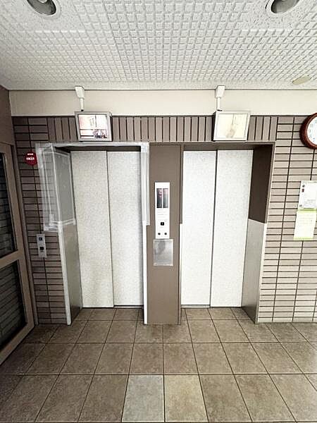 落ち着いた色合いのタイル床が特徴のエントランスです。二基のエレベーターが並びスムーズな移動が可能です。清潔感のある内装が印象的です。エントランス付近には掲示板や時計も設置されています。