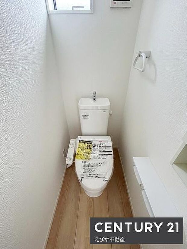 シンプルですっきりとしたトイレ空間です。壁紙とフローリングの組み合わせが印象的です。小窓から自然光が差し込み明るさを感じられます。ペーパーホルダーや収納棚も設置されています。清潔感ある仕上がりです。