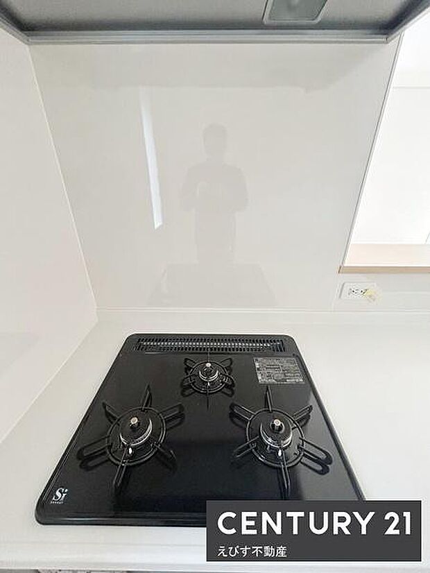 清潔感のあるキッチンでコンロ周りがスッキリとしています。三口ガスコンロが設置されており調理スペースが確保されています。天板や壁面は手入れがしやすい素材です。シンプルなデザインが特徴です。