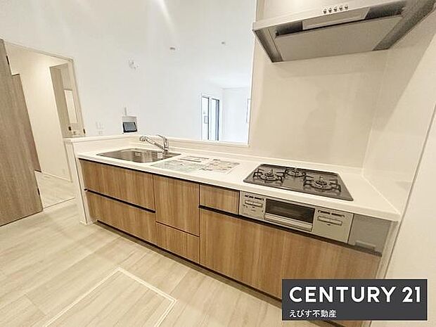 明るい空間に調和する木目調のキッチンが印象的です。ワークトップが広く使いやすい設計です。収納スペースも充実しています。ガスコンロや換気扇などの設備が揃っています。清潔感のある仕上がりです。