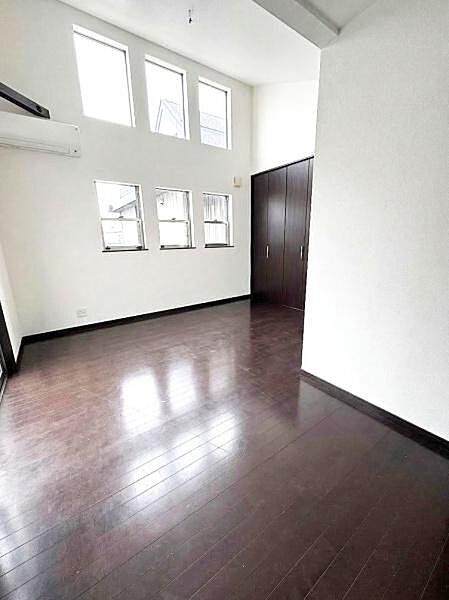 【一部AIによるモノ消し済み】洋室は天井が高く開放感があります。複数の窓からたっぷりと光が入り明るい空間です。ダークブラウンのフローリングが落ち着いた印象を与えます。収納スペースも備えられています。