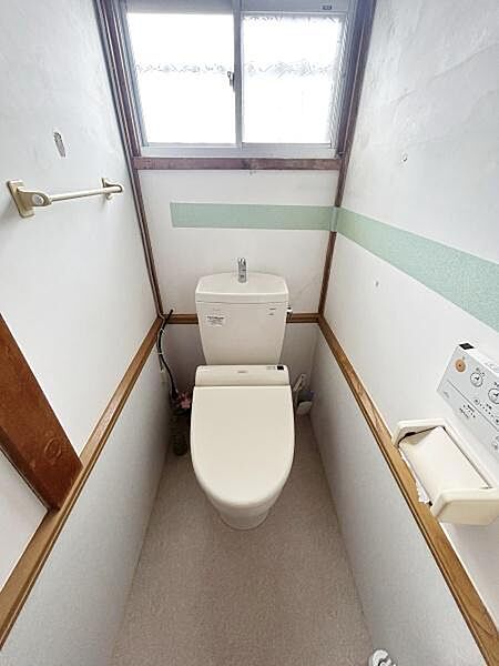 【一部AIによるモノ消し済み】窓付きでしっかり換気が可能なトイレです。壁面に手すりやペーパーホルダーも設置されています。シンプルな内装で清潔感があります。落ち着いた空間として利用できます。