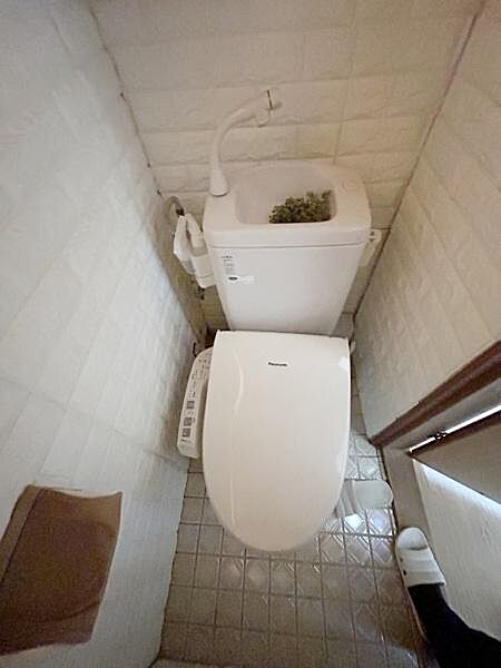 シンプルな内装のトイレでコンパクトな空間設計となっています。タンク上部には手洗い器が設置されています。ペーパーホルダーや操作パネルも使いやすい配置です。床はお手入れしやすいタイル仕様となっています。
