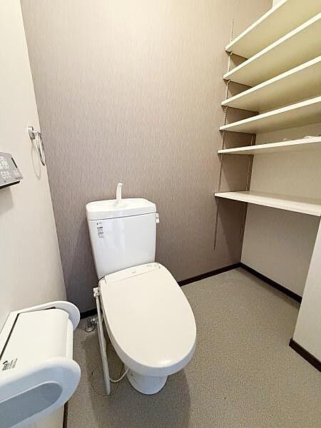 壁面に設置された収納棚が特徴のトイレです。清潔感のある床材と落ち着いた壁色で空間に統一感があります。手洗い付きのタンクが備わっており実用性も感じられます。