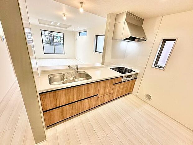 キッチンはシンプルなデザインで空間が広々と感じられます。木目調の収納が落ち着いた印象を与えています。十分な作業スペースが確保され窓からの光も取り入れられます。清潔感のある色調が魅力的です。