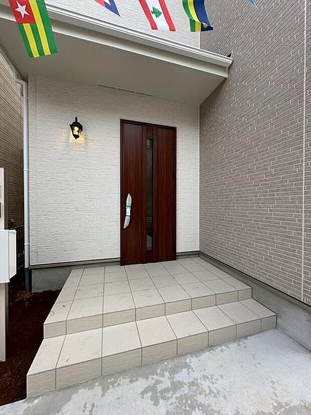 シンプルで清潔感のある玄関が印象的です。段差も緩やかで出入りしやすい設計になっています。玄関扉の木目が落ち着いた雰囲気を演出しています。外壁の明るい色合いが開放感を与えます。