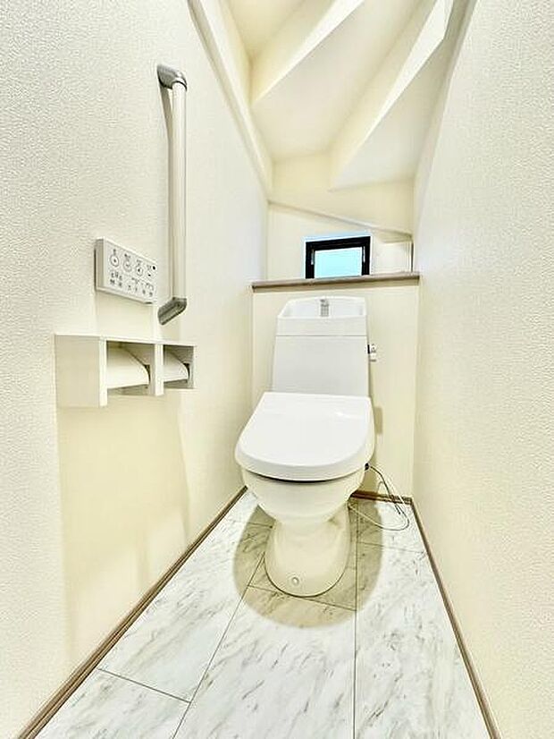 明るい光が差し込むトイレ空間です。壁や床は清潔感のある色調でまとめられています。使いやすい手すりや操作パネルも設置されています。収納棚も付いており便利な設備が整っています。