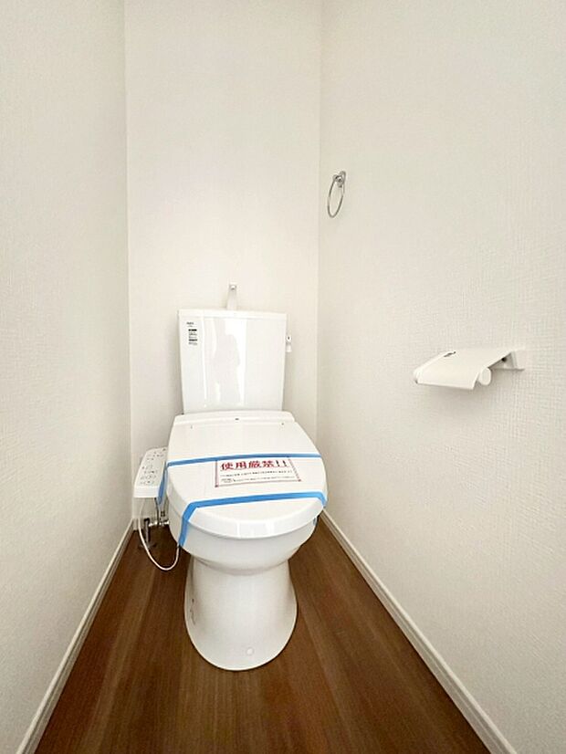 白を基調とした清潔感のあるトイレです。すっきりとしたデザインで空間が広く感じられます。床材が木目調で落ち着きのある印象を与えます。ペーパーホルダーやタオルリングも設置されています。