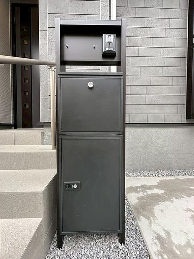 シンプルで機能的なデザインの宅配ボックスです。玄関先に設置されており周囲の建物とも調和しています。グレーの外壁に合ったカラーリングが特徴的です。置き場所もスペースを有効活用できる仕様となっています。