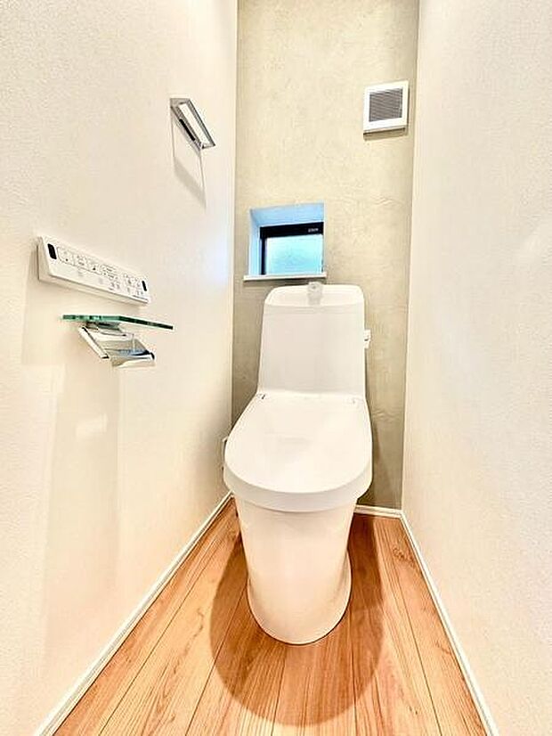 清潔感のあるトイレ空間です。小窓が設けられており換気がしやすくなっています。壁付けのリモコンで操作も簡単です。床は木目調で温かみを感じられます。コンパクトにまとまった設計です。