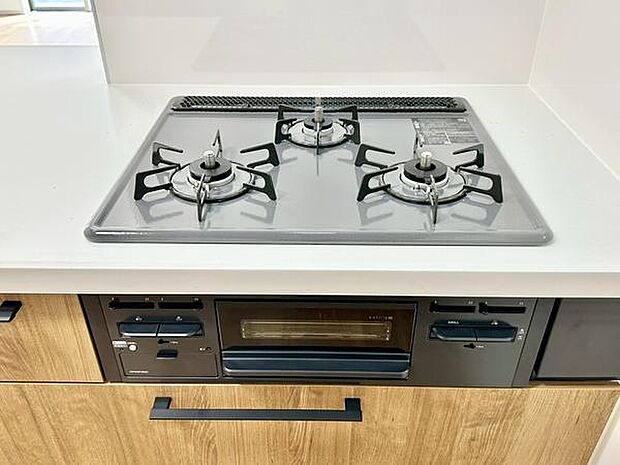 使いやすい三口コンロが設置されていますので調理の幅が広がります。グリルも付いておりさまざまな料理に対応できます。木目調の収納とブラックの取っ手が調和し全体的に落ち着いた印象のキッチンです。