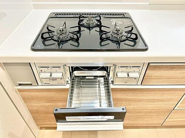 3口のコンロが搭載されたキッチンです。グリルも備わっており幅広い調理に活用できます。シンプルですっきりとしたデザインが印象的です。木目調の扉が空間に温かみをプラスします。