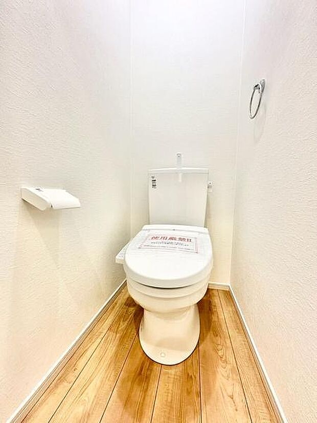 明るい色合いの床材が特徴的なトイレです。照明がしっかりしており空間がすっきりとしています。シンプルなデザインで清潔感があります。壁面にはペーパーホルダーやタオルリングも備わっています。