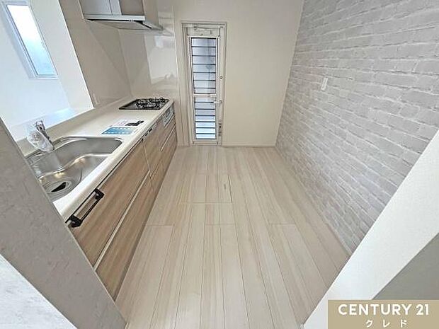 レンガ調のアクセントクロスが印象的なキッチンスペース。背面にはカップボードを置いてもゆとりがあり、すっきりと使えます。勝手口付きで明るく、換気やゴミ出しにも便利な動線です。