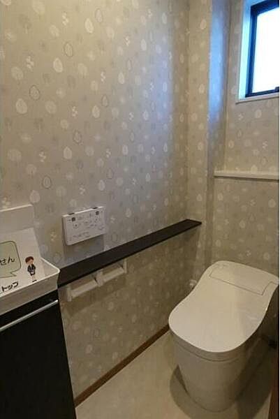 窓が設置されており明るさと換気に配慮されたトイレです。壁紙のデザインが空間にやわらかな印象を与えています。使いやすい位置にペーパーホルダーや棚が設置されています。清潔感を感じる内装です。
