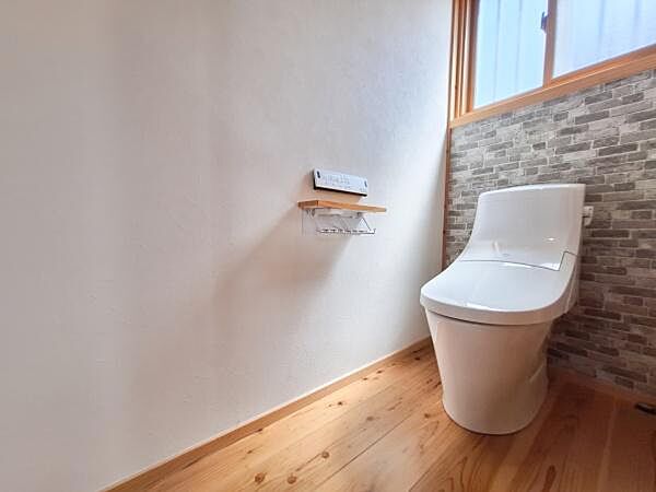 清潔感のあるトイレ空間です。木目調の床が温かみを演出しています。窓があり自然光が差し込む明るい印象です。収納や小物置きにも配慮された設計です。