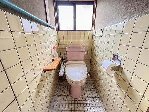明るい自然光が差し込む窓付きのトイレです。壁と床のタイルが清潔感を演出しています。収納やペーパー棚も設けられています。温水洗浄便座が設置されています。小物置きスペースも便利です。