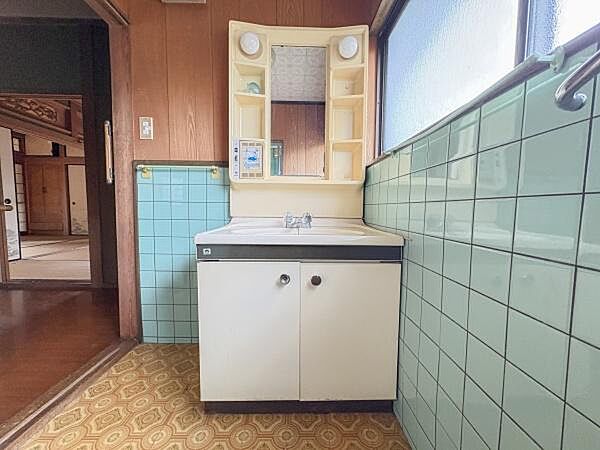 洗面台には使いやすい収納スペースが備わっています。明るい光が差し込む窓際に設置されています。清潔感を感じるタイル張りの壁が特徴です。鏡の両脇には便利な小物置きスペースがあります。